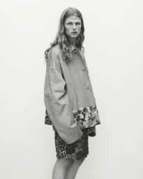 Model with long wavy hair posing in an oversized buttoned jacket and textured shorts against a seamless studio backdrop.