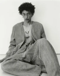 Model wearing an open textured suit with drawstring trousers and a pendant necklace, seated against a white background.