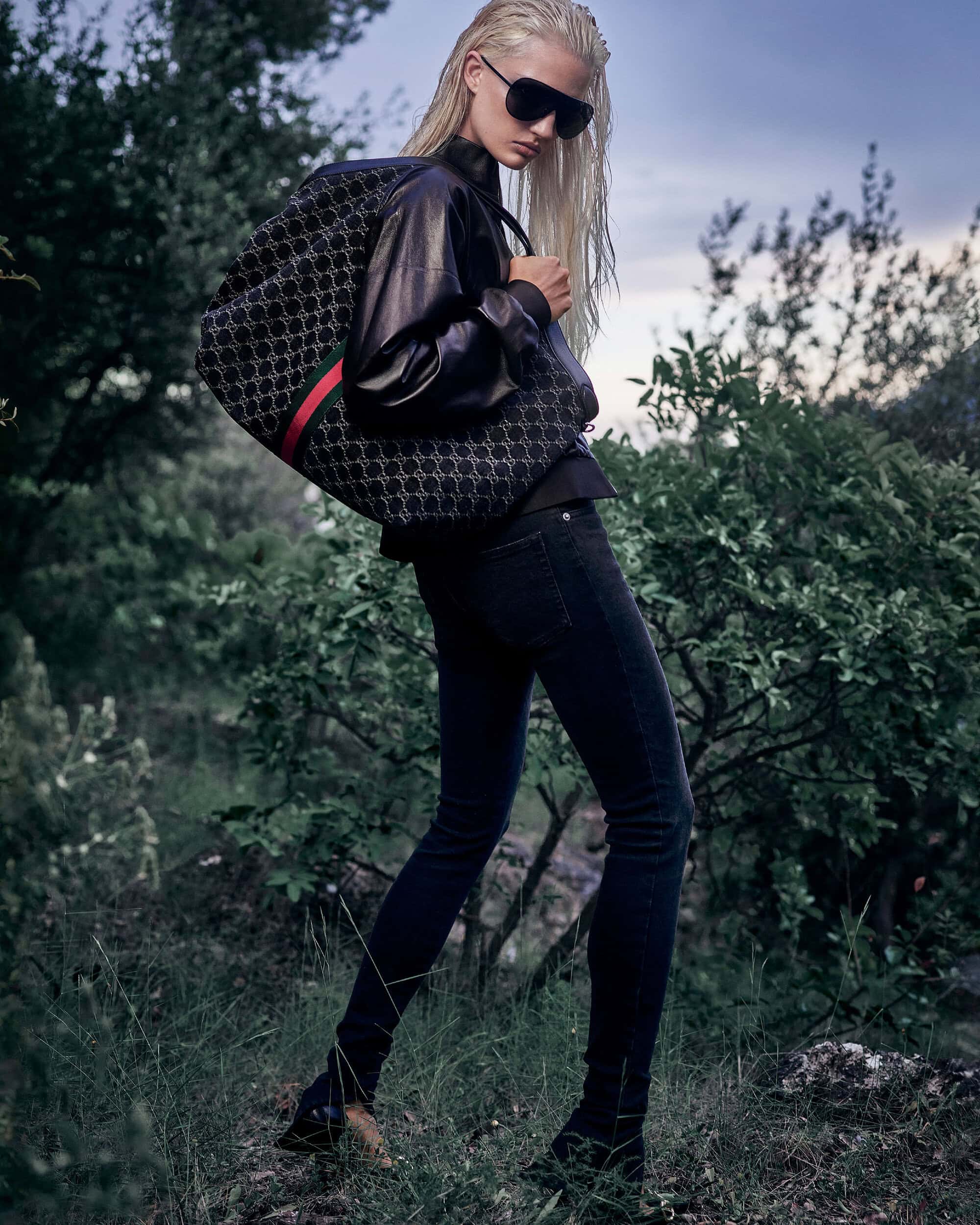 Blonde model in sunglasses and a black leather jacket carrying a large monogram Gucci tote bag in a wooded outdoor setting.