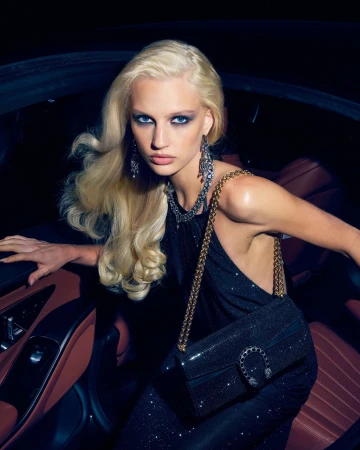 Blonde woman in a sparkling black halter dress posing inside a luxury car holding a black Gucci Dionysus chain bag.