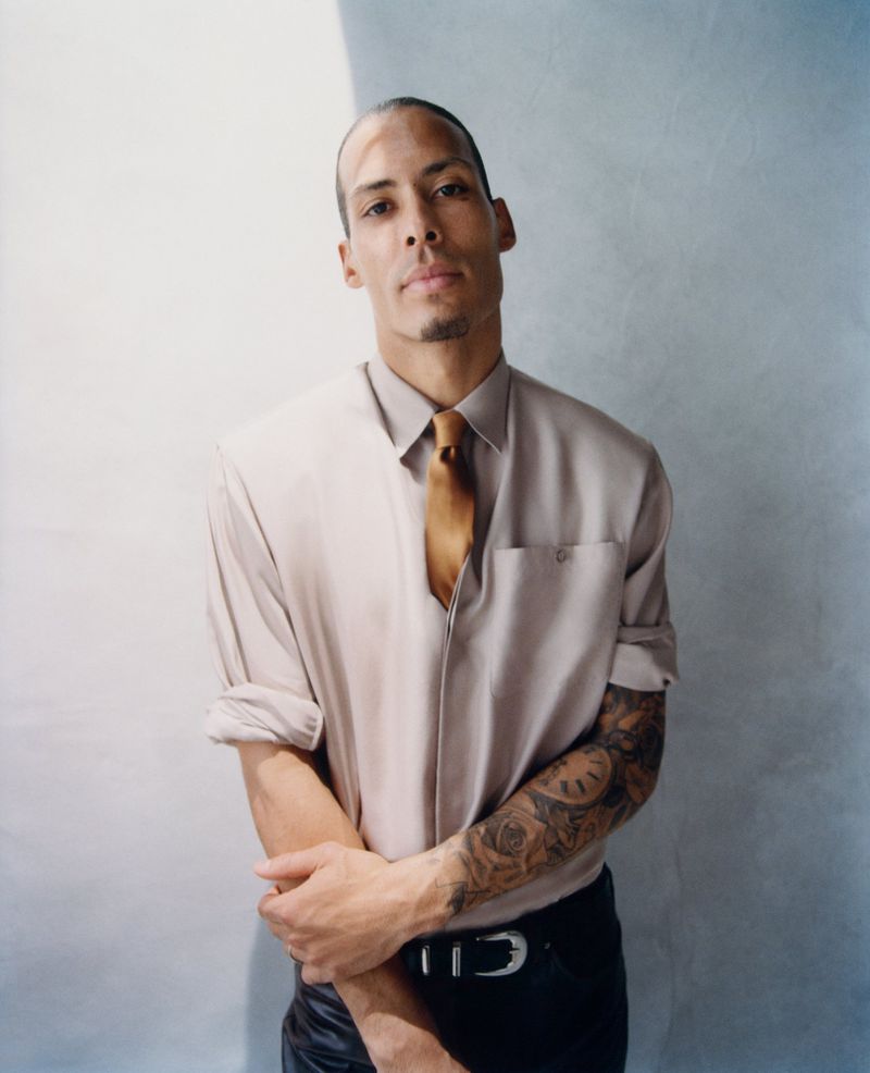 Virgil van Dijk wearing a beige rolled-sleeve collared shirt with a gold necktie and black pants against a soft studio backdrop.