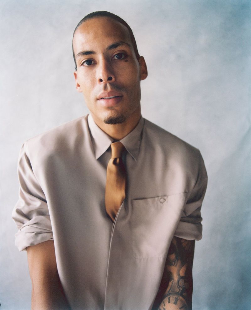 Virgil van Dijk wearing a beige collared shirt and golden brown tie against a textured studio backdrop.