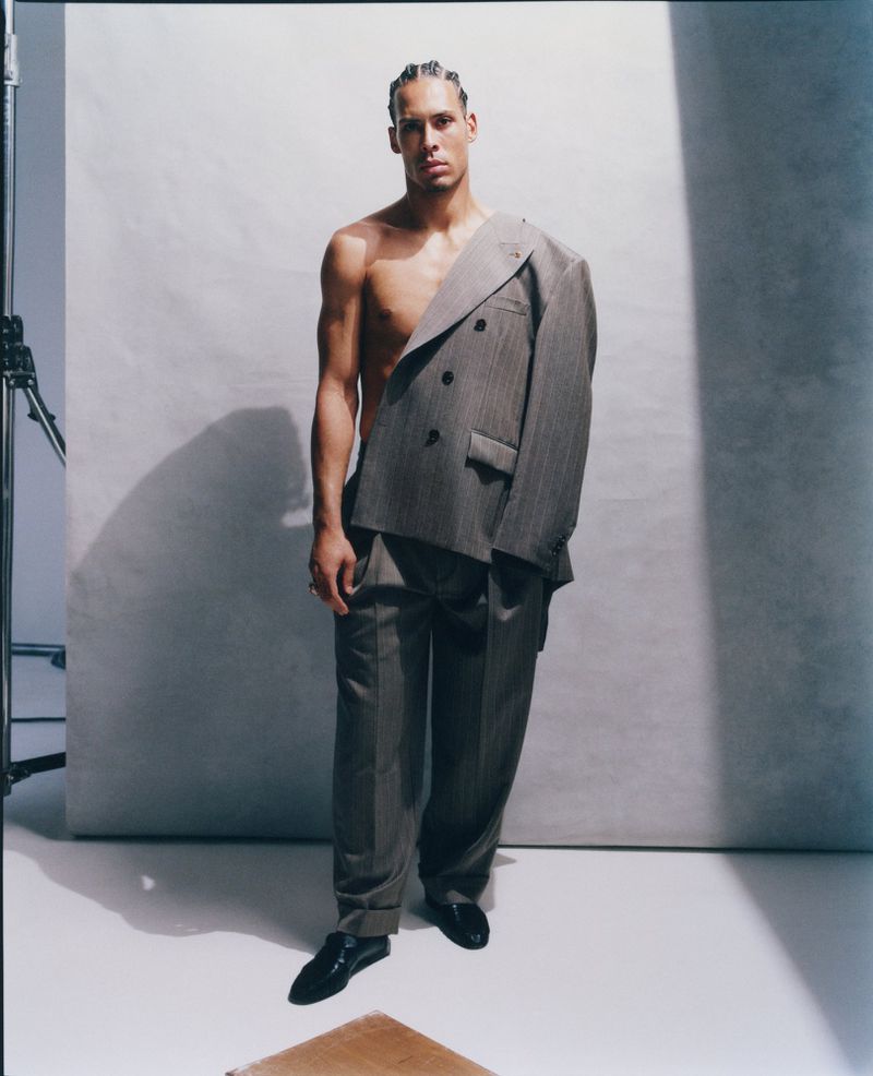 Virgil van Dijk standing shirtless wearing an asymmetrical grey tailored blazer and matching trousers in a studio setting.