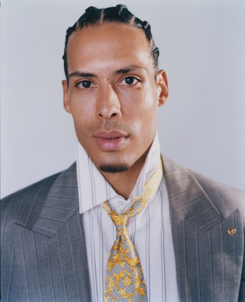 Portrait of athlete Virgil van Dijk wearing a grey blazer, striped shirt, and patterned gold tie against a plain light background