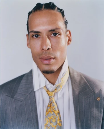 Portrait of athlete Virgil van Dijk wearing a grey blazer, striped shirt, and patterned gold tie against a plain light background