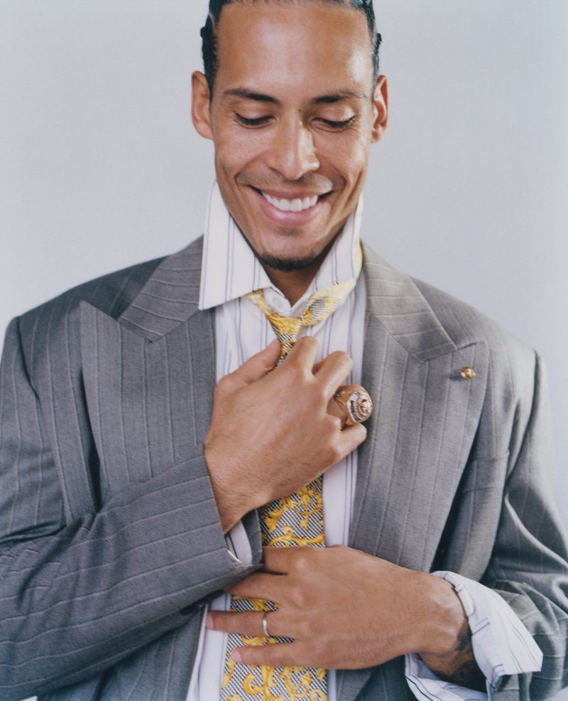 Virgil van Dijk smiling while adjusting a yellow patterned tie and wearing a grey pinstripe suit against a studio backdrop.