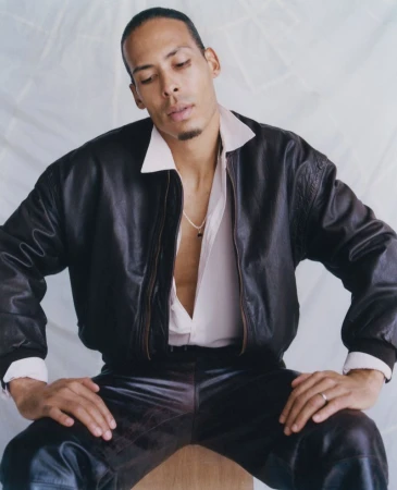 Virgil van Dijk seated in a dark leather bomber jacket and unbuttoned pale shirt against a textured backdrop.