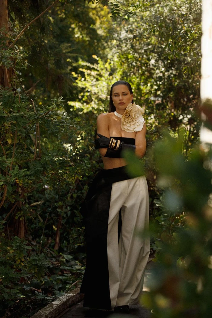 Adriana Lima poses outdoors in a black strapless top, wide-leg trousers, long black gloves, and a large white floral neckpiece.