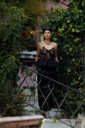 Adriana Lima wearing a black lace tiered top and headpiece stands on outdoor steps framed by ivy and a metal railing.