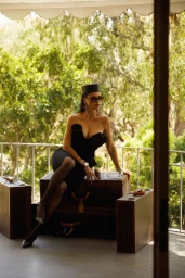 Adriana Lima seated on vintage luggage wearing a black strapless dress, pillbox hat, and sunglasses on an outdoor balcony.