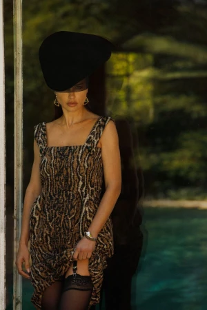 Adriana Lima wearing a sculpted black hat with veil, leopard textured dress, and stockings by a glass window.