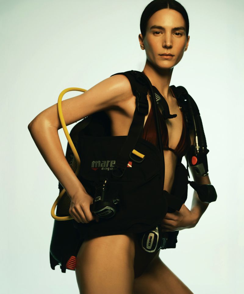 Model Mijo Mihaljcic posing in a scuba diving BCD vest and harness over swimwear against a pale studio background.