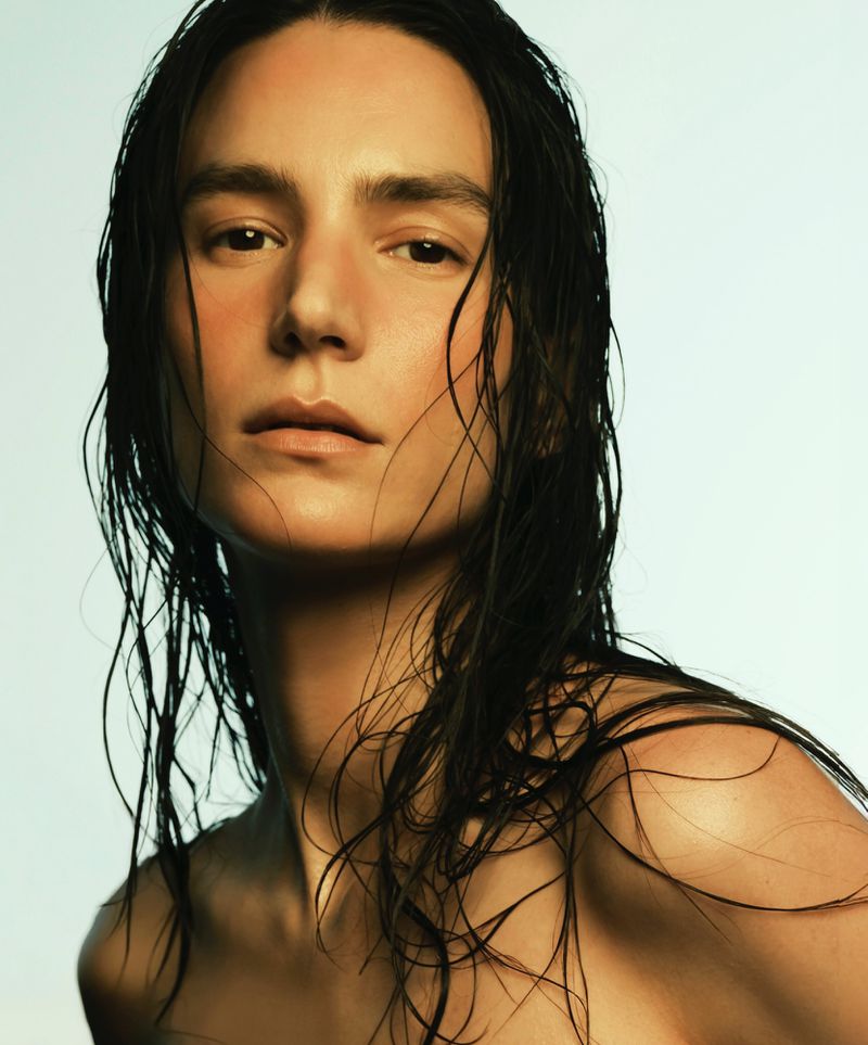 Close-up portrait of model Mijo Mihaljcic with wet dark hair and bare shoulders against a pale backdrop.