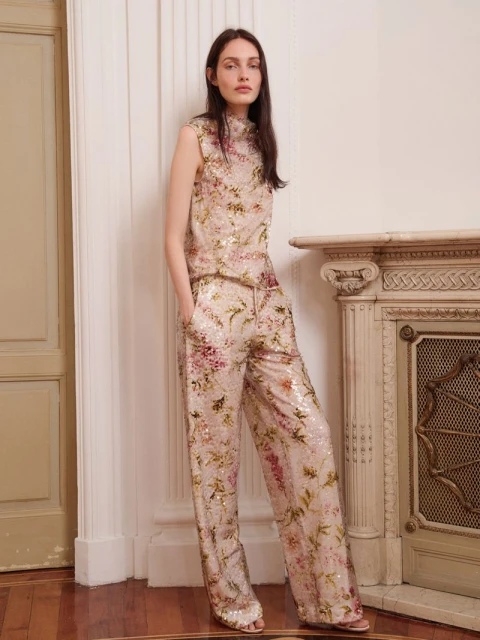 A woman models a floral-patterned sleeveless top and wide-leg pants in a fashion shoot. She stands indoors with a neutral expression.