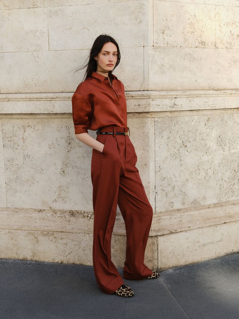 Brunette fashion model in rust-colored blouse and trousers poses against a stone wall in natural light showcasing editorial fashion.
