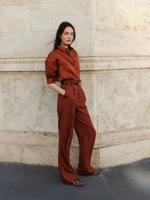 Brunette fashion model in rust-colored blouse and trousers poses against a stone wall in natural light showcasing editorial fashion.