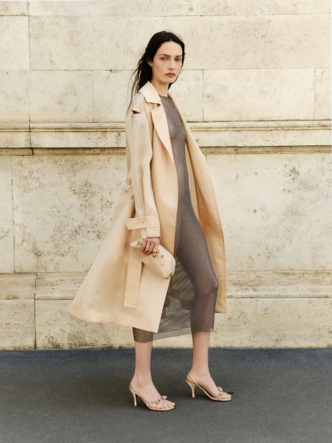 A slender woman with long dark hair models a beige trench coat, gray mesh dress, and matching heeled sandals outdoors against a textured wall.