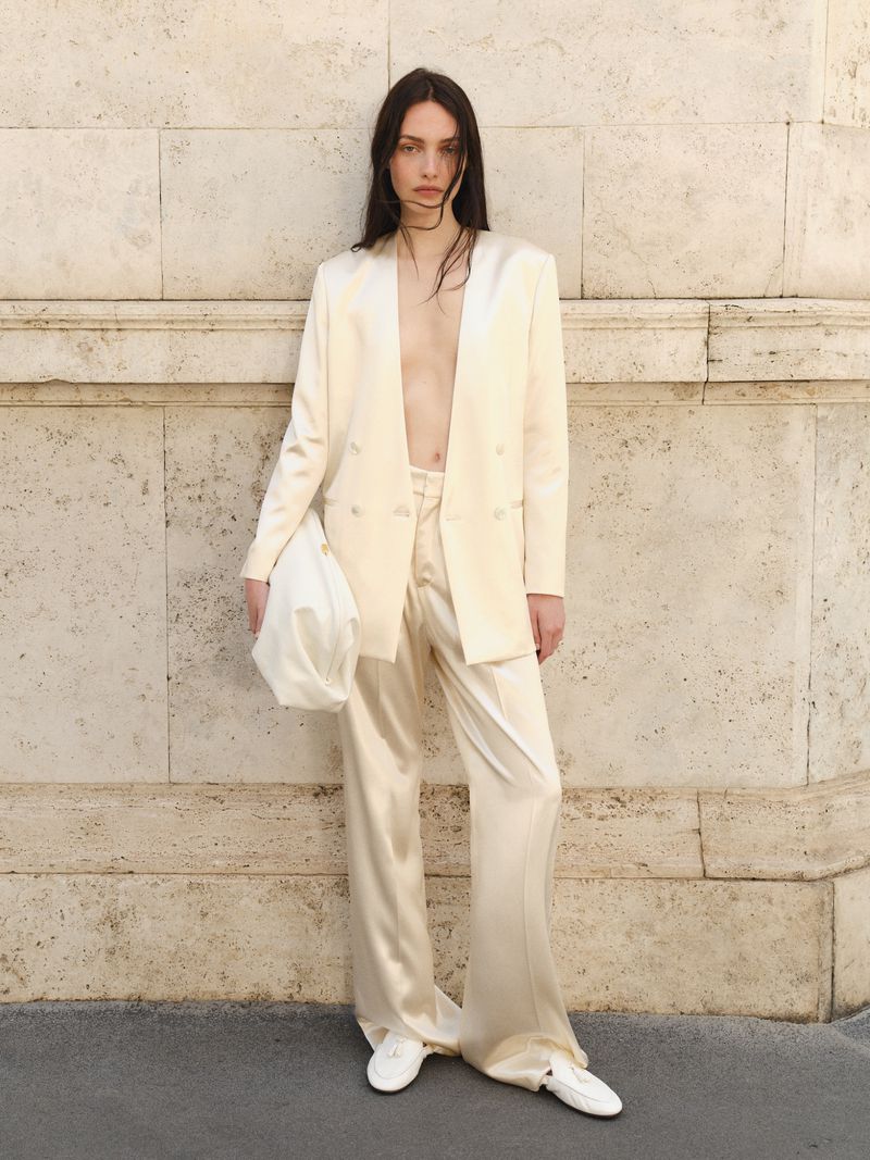 A slender fashion model poses against a neutral wall, wearing an off-white satin suit, white sneakers, and carrying a matching handbag.