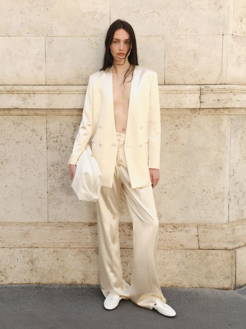 A slender fashion model poses against a neutral wall, wearing an off-white satin suit, white sneakers, and carrying a matching handbag.