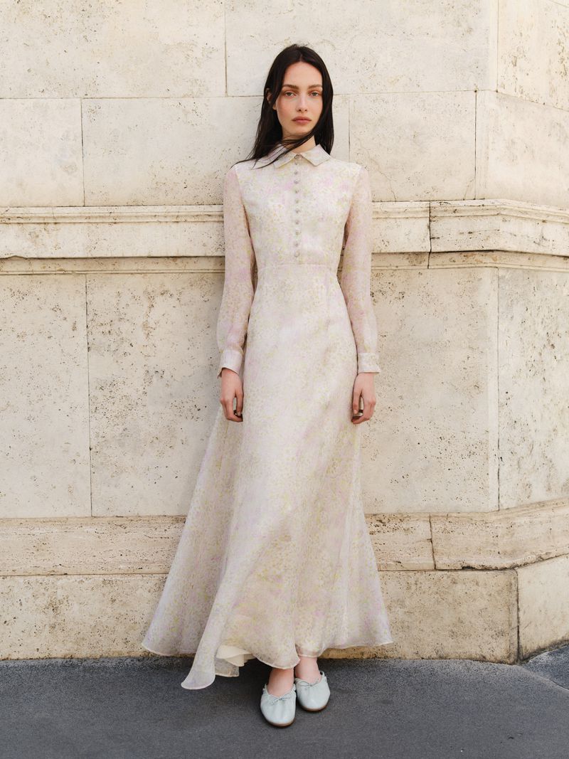 A slender woman poses outdoors in a long-sleeved, light-colored dress with a buttoned bodice, complemented by matching flats.