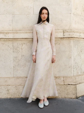 A slender woman poses outdoors in a long-sleeved, light-colored dress with a buttoned bodice, complemented by matching flats.