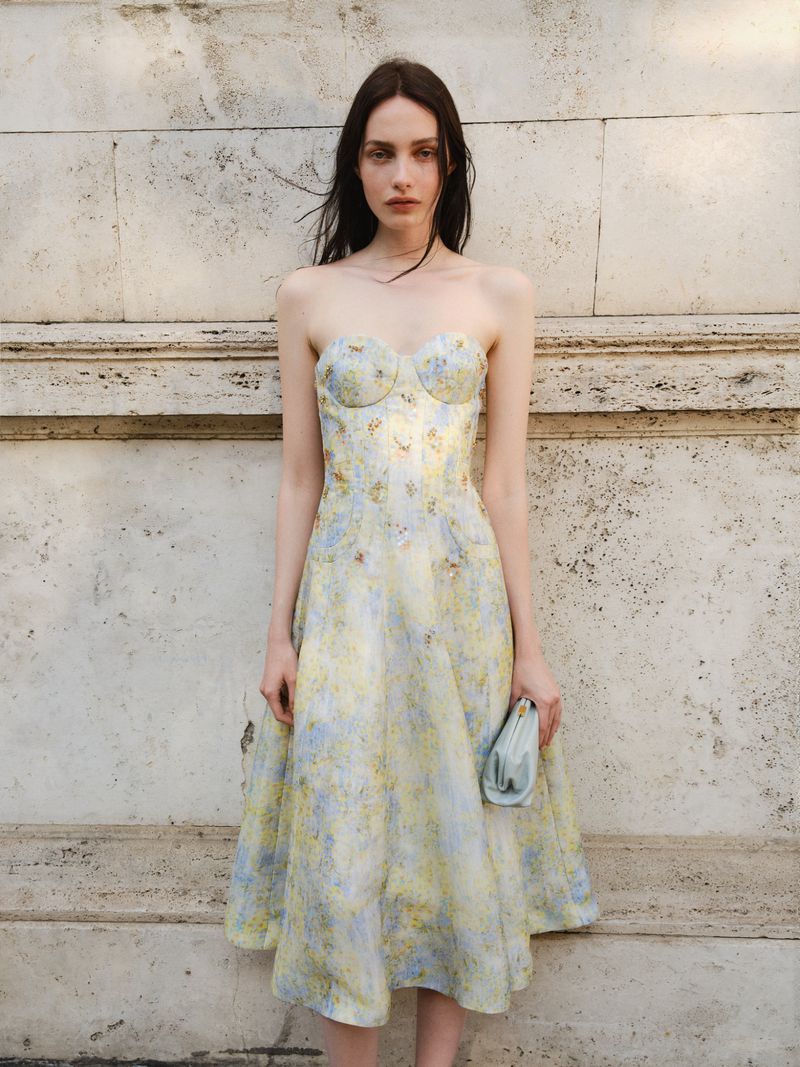 Elegant woman in a floral dress and clutch poses against a stone wall in natural light, capturing a blend of classic style and beauty.
