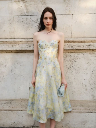 Elegant woman in a floral dress and clutch poses against a stone wall in natural light, capturing a blend of classic style and beauty.