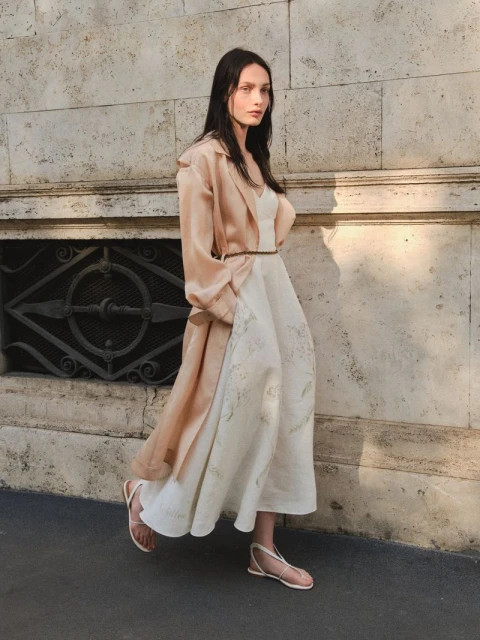 Elegant woman in a light-colored dress and coat walks by a stone building with metal window grates in an outdoor fashion setting.
