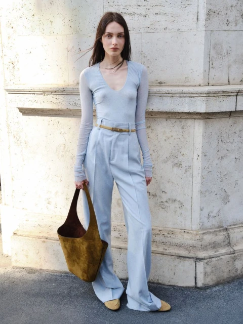 Fashionable woman in a light blue outfit poses with a brown tote bag in an urban setting against a textured stone wall.