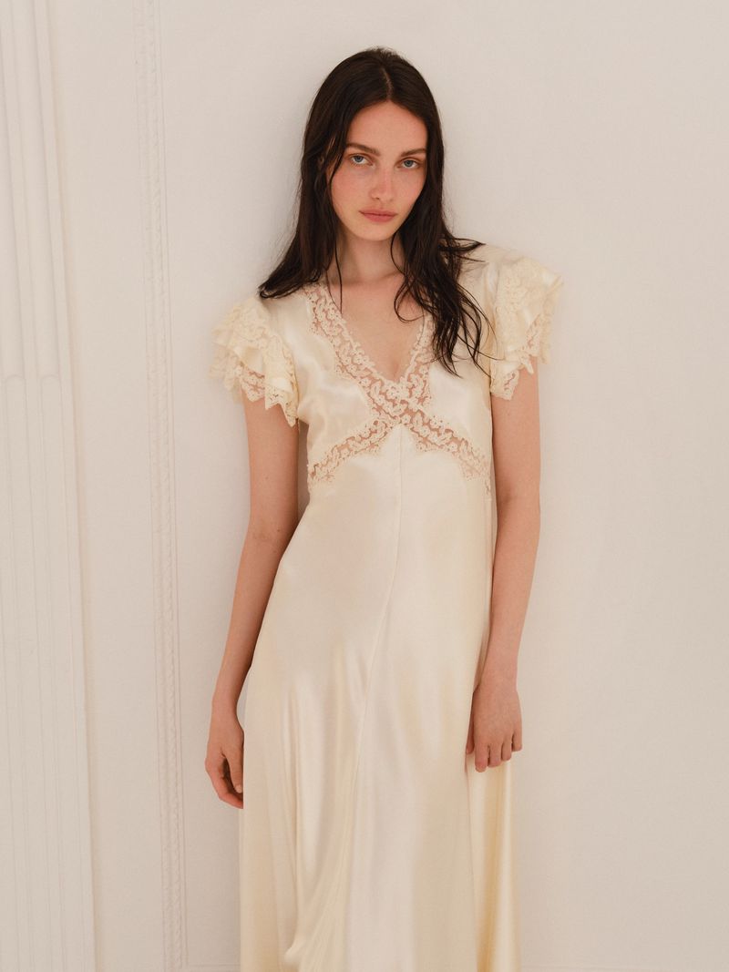 Elegant woman poses in a pale yellow vintage gown with lace details against a neutral background, showcasing fashion and style.
