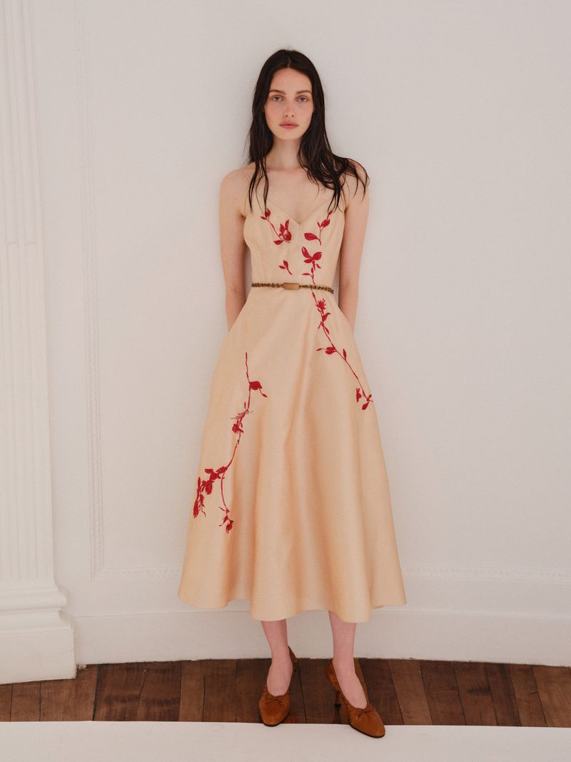 Full-length shot of a brunette woman in a peach-colored dress with floral embroidery, wearing brown shoes in an indoor setting.