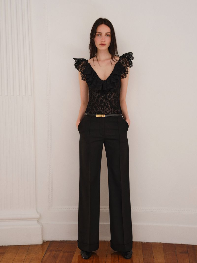 Full-length portrait of a young adult woman in a black lace top and black pants standing indoors near a white wall and column.