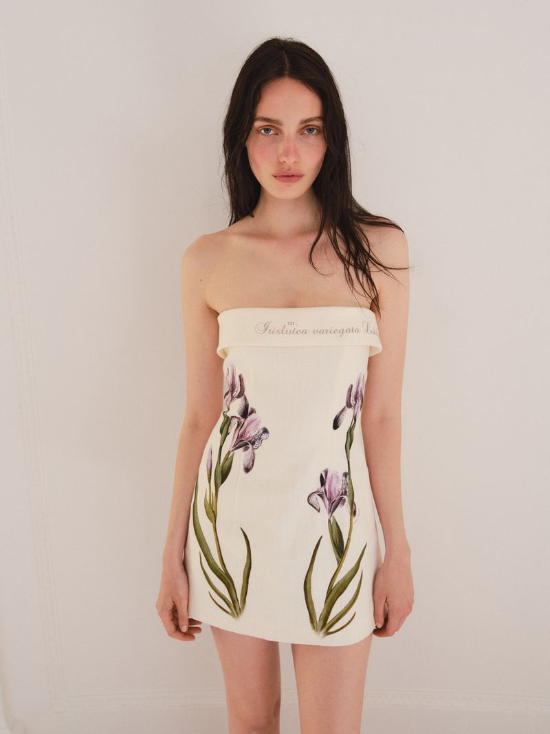 Brunette woman in a strapless, cream mini dress with painted iris flowers posing against a white background in a studio setting.