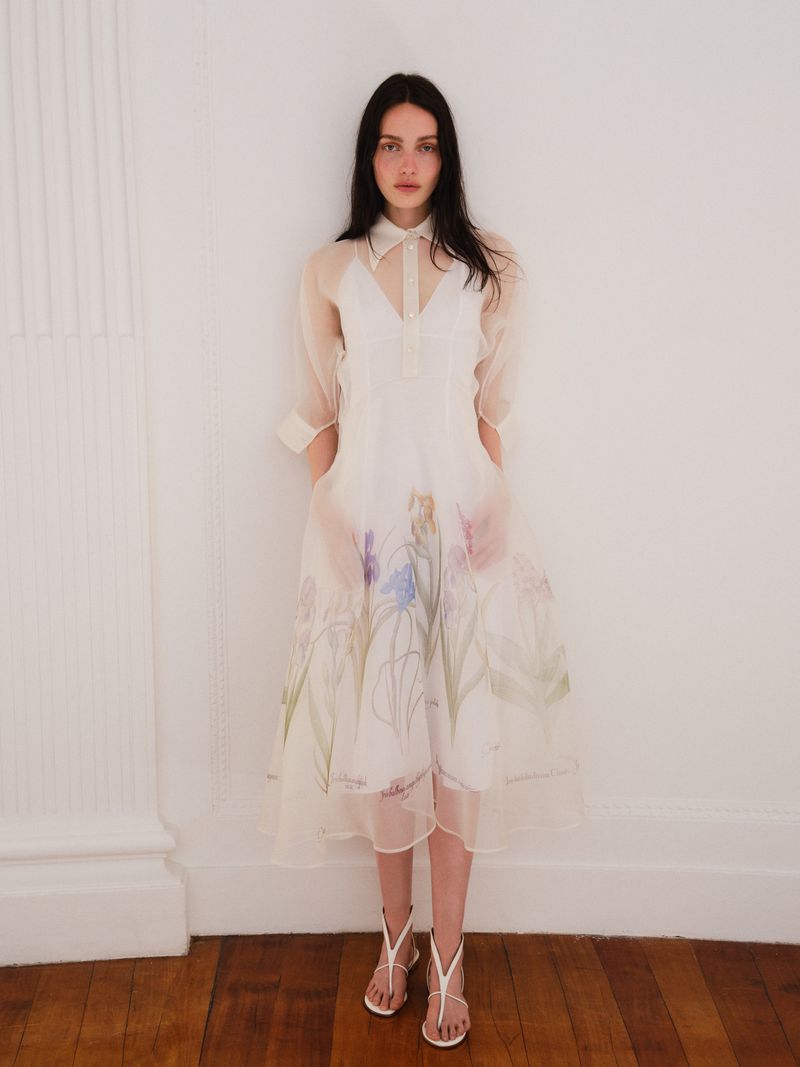 A woman in a semi-sheer white dress with floral details poses against a white wall and wood floor in a fashion editorial shot.