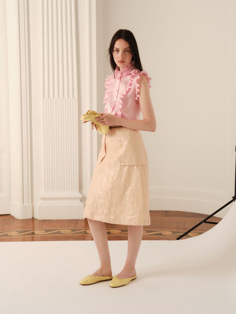 A young adult woman poses in a light pink ruffled top and light beige skirt with yellow flats in a photo studio with neutral tones.