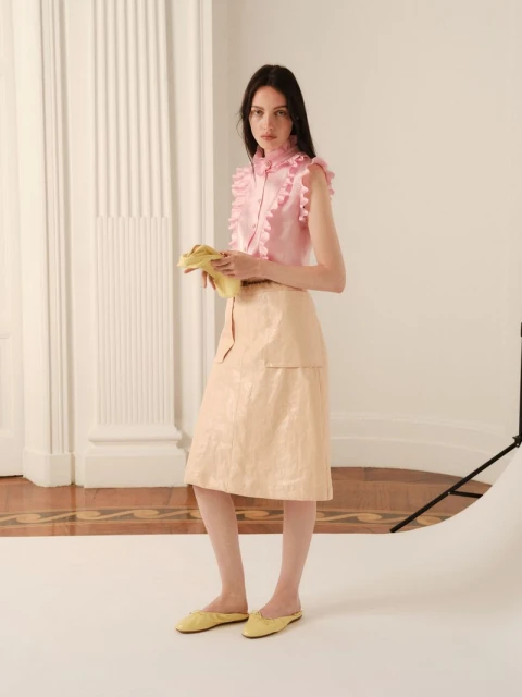 A young adult woman poses in a light pink ruffled top and light beige skirt with yellow flats in a photo studio with neutral tones.