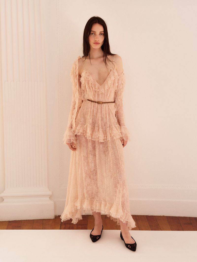 Elegant woman in a ruffled, sheer peach maxi dress poses in a bright photo studio with a column against a white backdrop.