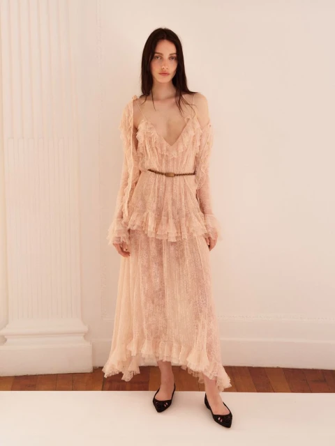 Elegant woman in a ruffled, sheer peach maxi dress poses in a bright photo studio with a column against a white backdrop.