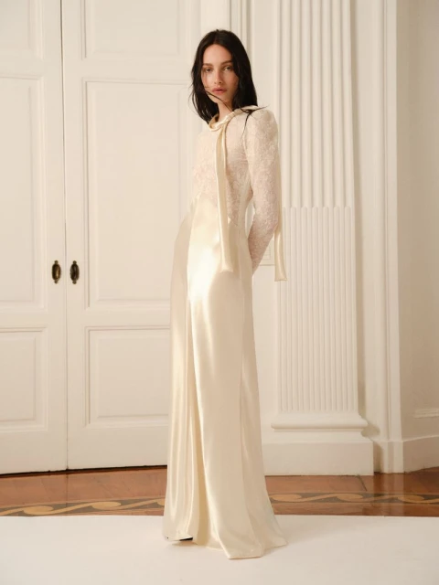 A woman in a long, off-white dress stands in front of white doors. The dress has lace sleeves and a tie at the neck.