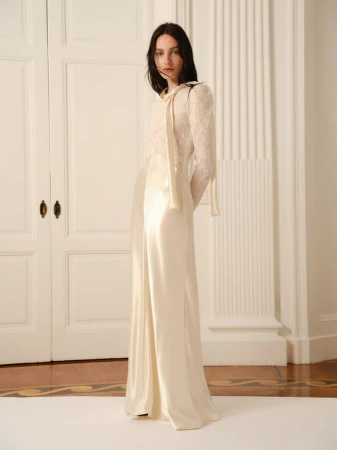 A woman in a long, off-white dress stands in front of white doors. The dress has lace sleeves and a tie at the neck.