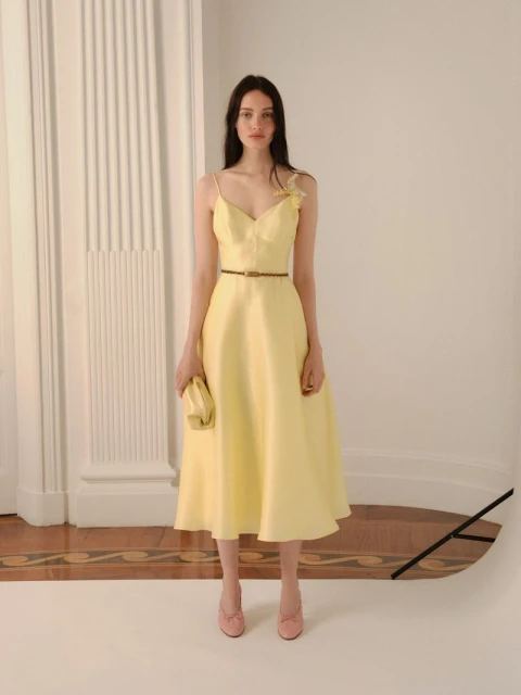 Elegant woman in a pale yellow midi dress with a brown belt, holding a matching clutch, standing indoors with a neutral backdrop.