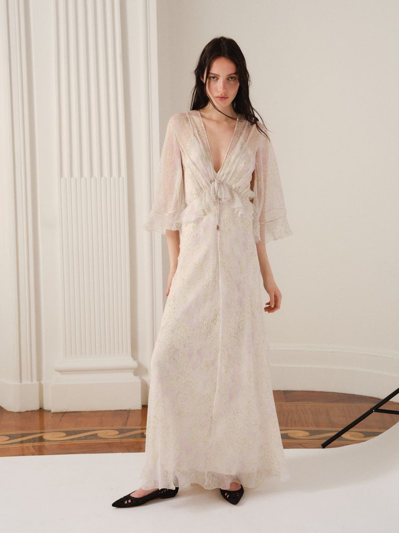 A young adult woman poses indoors wearing an off-white, floor-length dress with transparent sleeves and black flats, gazing at the camera.