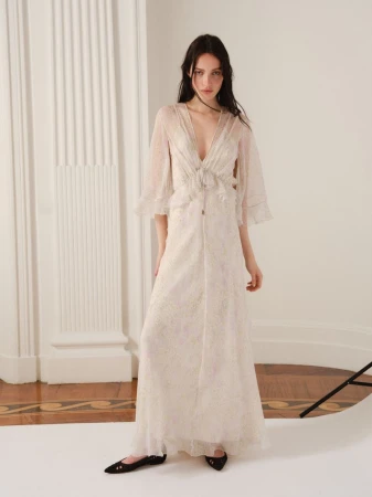 A young adult woman poses indoors wearing an off-white, floor-length dress with transparent sleeves and black flats, gazing at the camera.