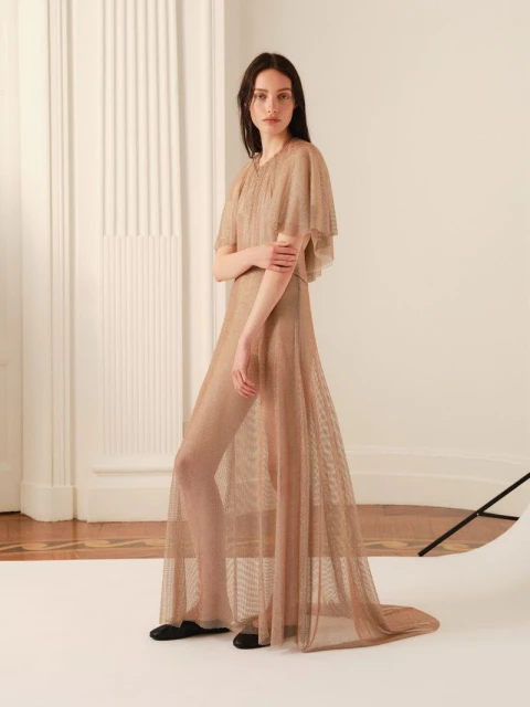 Fashion shot of a slender brunette woman posing in a light brown, floor-length sheer gown with short cape sleeves in a bright studio.