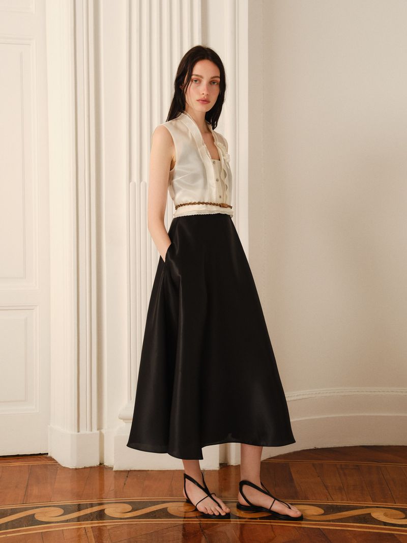 A woman models a cream-colored sleeveless top and black skirt indoors, with hands in pockets, on a wooden floor against a neutral wall.