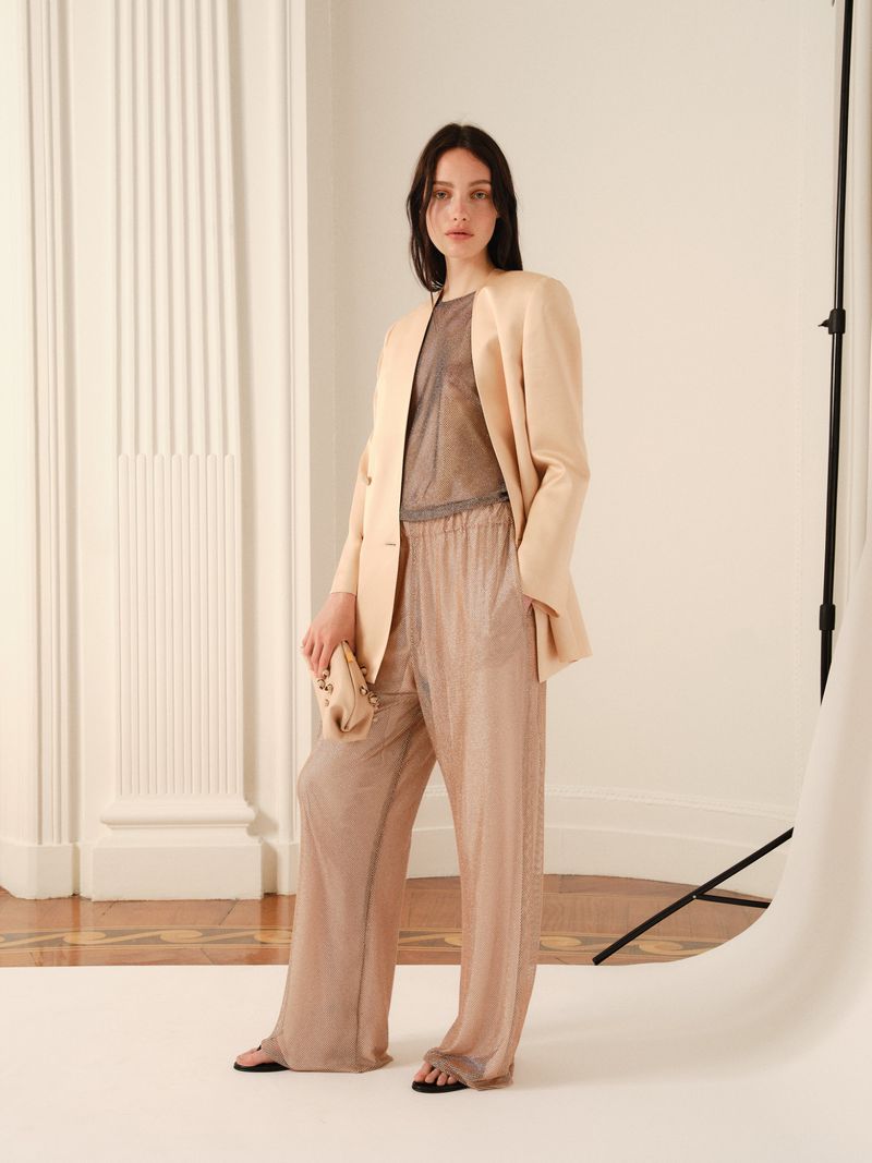 A young adult female model poses indoors in a light-beige blazer, matching loose-fitting trousers, and a metallic top while holding a clutch.