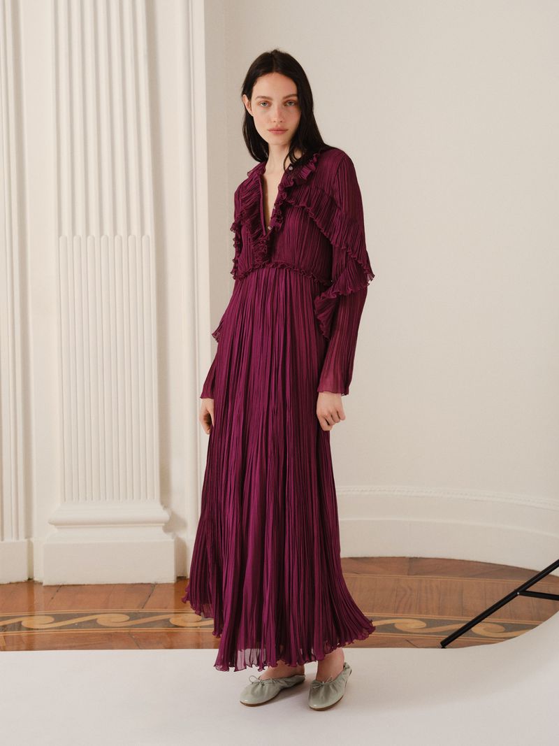 Elegant woman in a long-sleeve, floor-length magenta dress with ruffles, posing indoors with classic architecture columns.