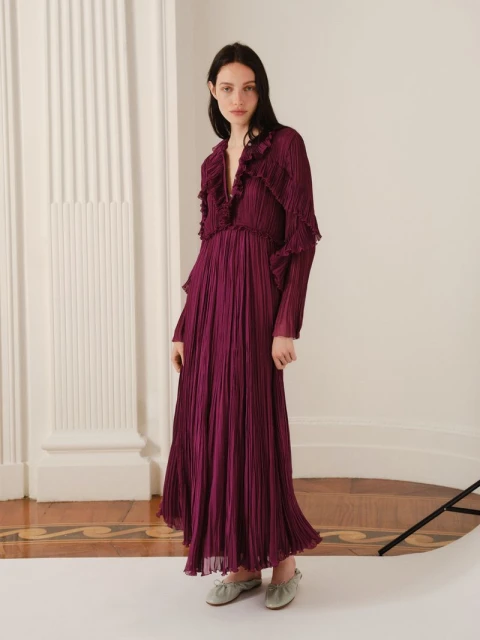 Elegant woman in a long-sleeve, floor-length magenta dress with ruffles, posing indoors with classic architecture columns.