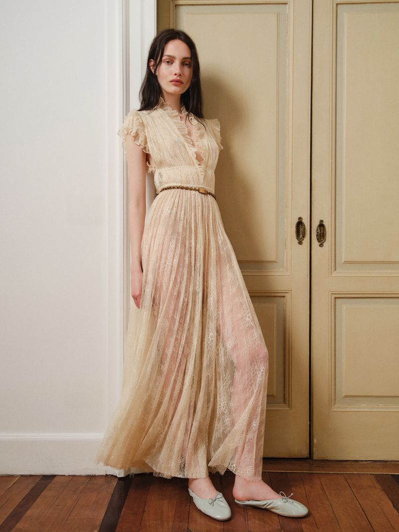 A woman in a long, light-colored dress poses in an indoor setting with hardwood floors and paneled doors for a fashion shoot.