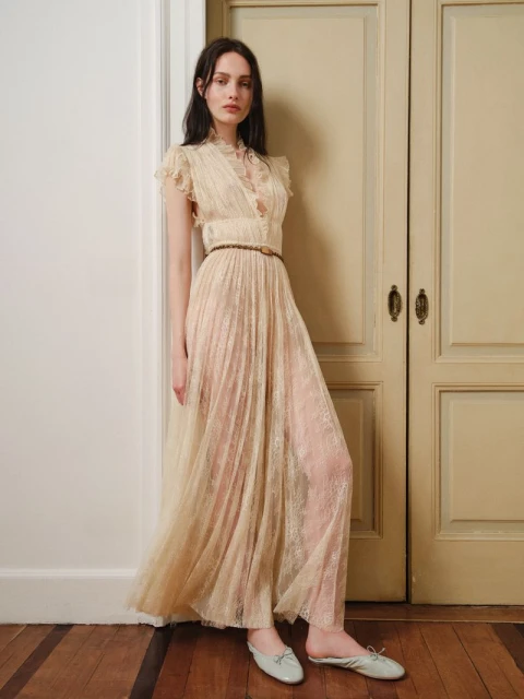 A woman in a long, light-colored dress poses in an indoor setting with hardwood floors and paneled doors for a fashion shoot.
